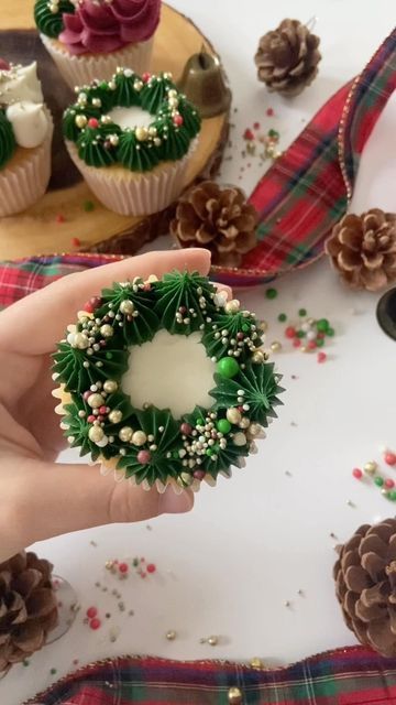 a hand holding a cupcake decorated with green and white frosting, surrounded by pine cones