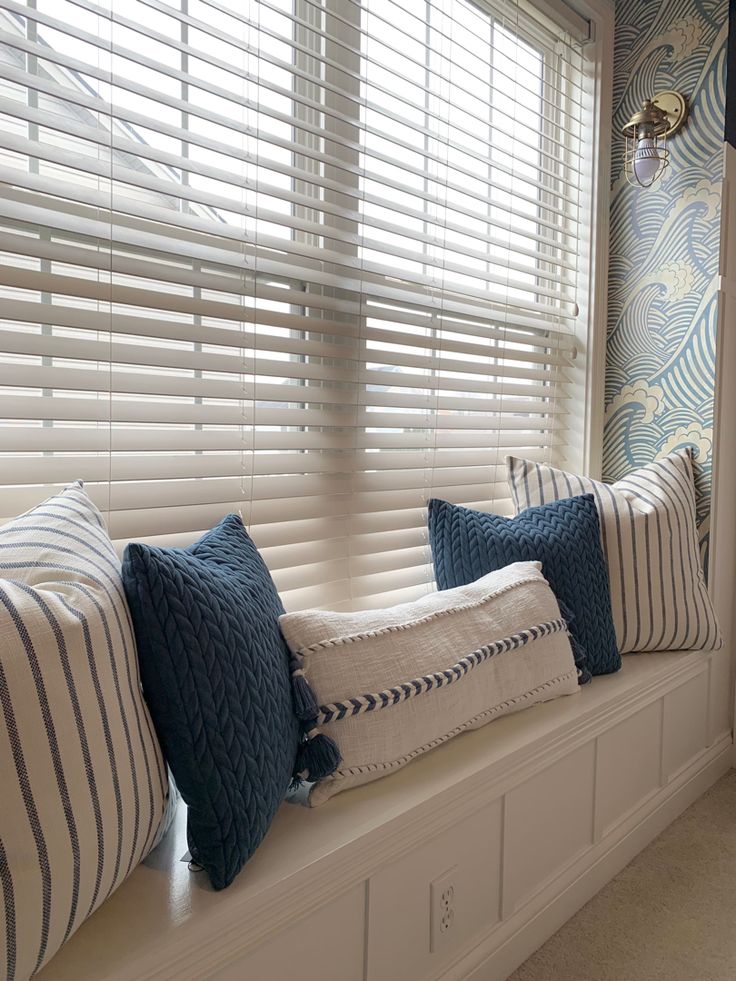 a window seat with several pillows on it in front of a window covered by blinds