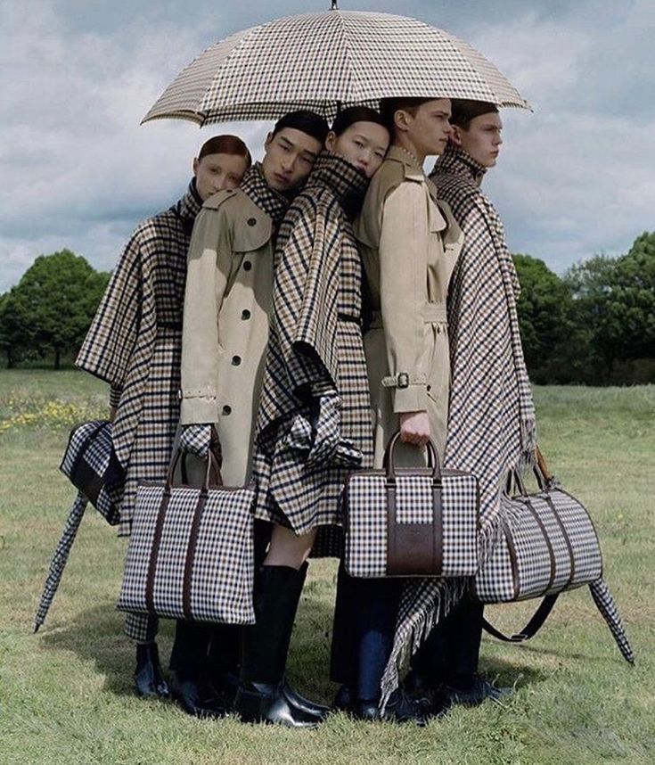 four men in trench coats are standing under an umbrella with their hands on their hipss