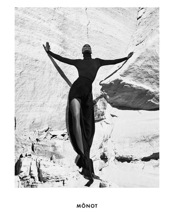 a black and white photo of a woman standing in front of a rock formation with her arms stretched out