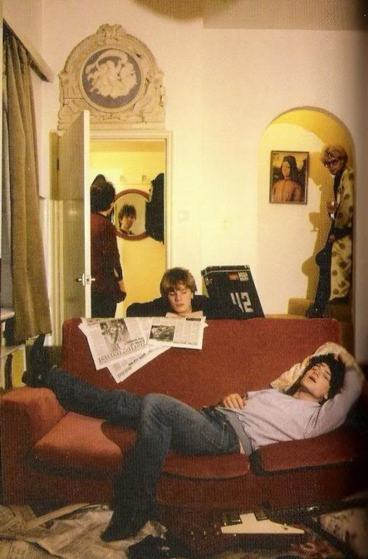 two people laying on a red couch in a living room next to a guitar and bookshelf