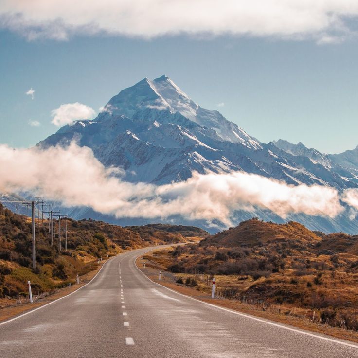 an empty road in front of a large mountain with snow on the top and clouds flying over it