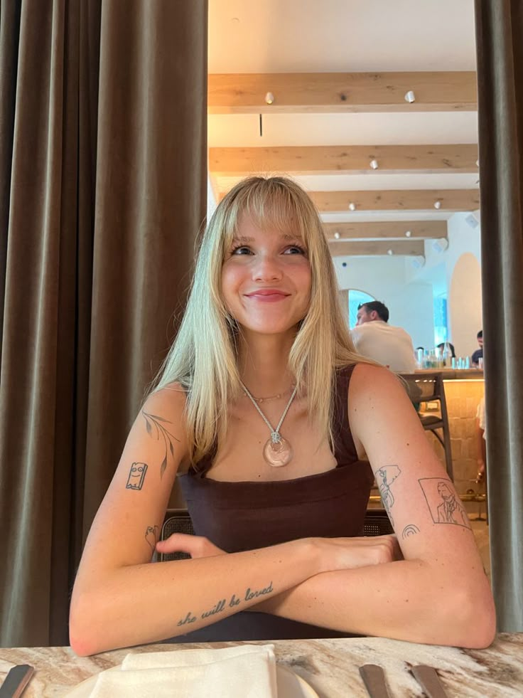 a woman sitting at a table with her arms crossed and tattoos on her chest, smiling