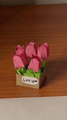 some pink flowers are sitting in a small box on a wooden table with the word luv u written on it