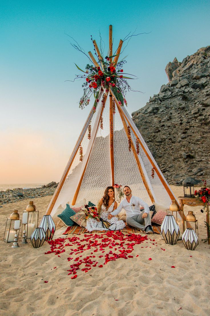 two people sitting in front of a teepee on the beach with rose petals all over it