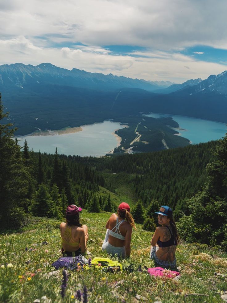 three women sitting on the side of a mountain looking at a lake and mountains in the distance