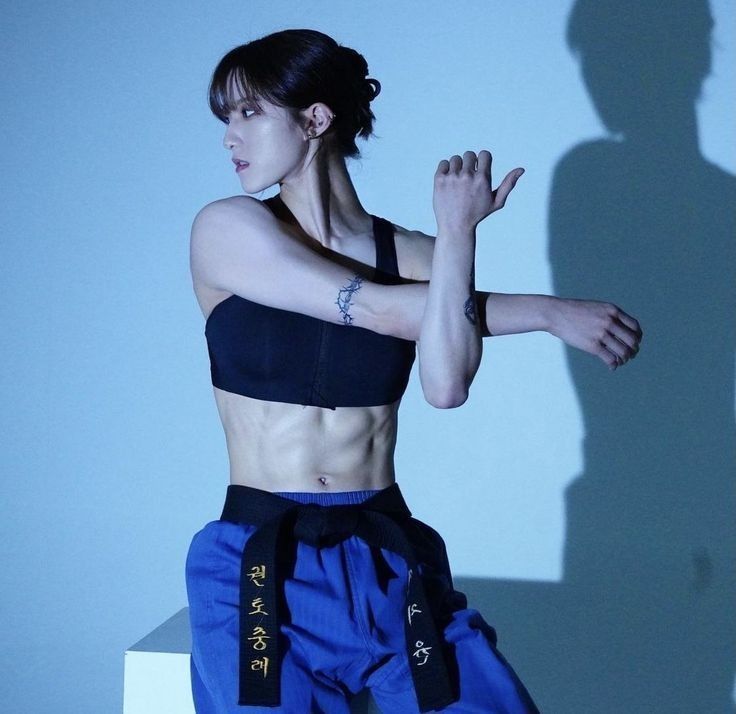 a woman standing in front of a white wall wearing blue pants and a black top