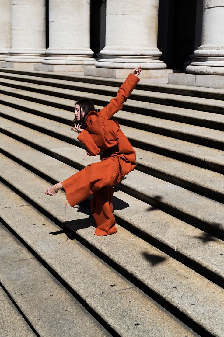 a woman in an orange outfit is dancing on some steps with her hand up to the side