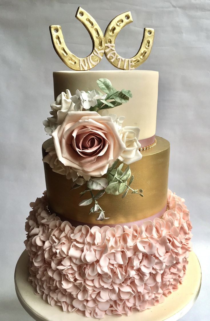 a three tiered cake with pink flowers and gold letters on top, sitting on a white pedestal