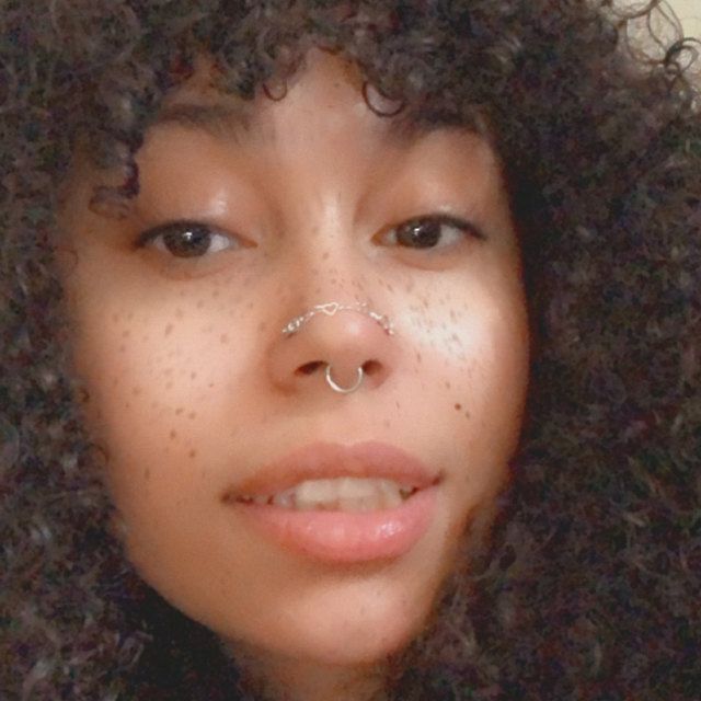 a close up of a person with curly hair and piercings on her nose, looking at the camera