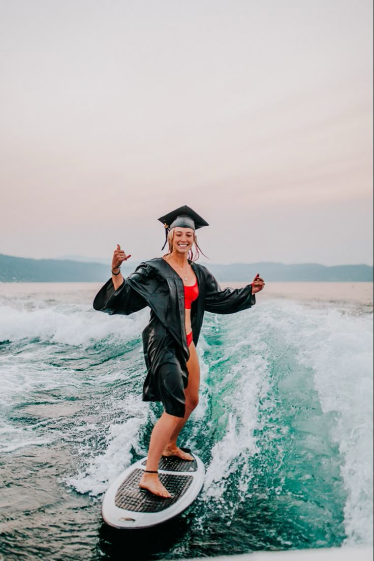 a woman in a graduation gown riding a surfboard on the ocean with waves behind her