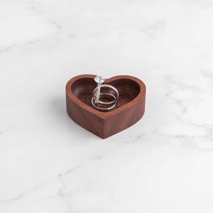 a wooden heart shaped box with two wedding rings in it sitting on a marble surface