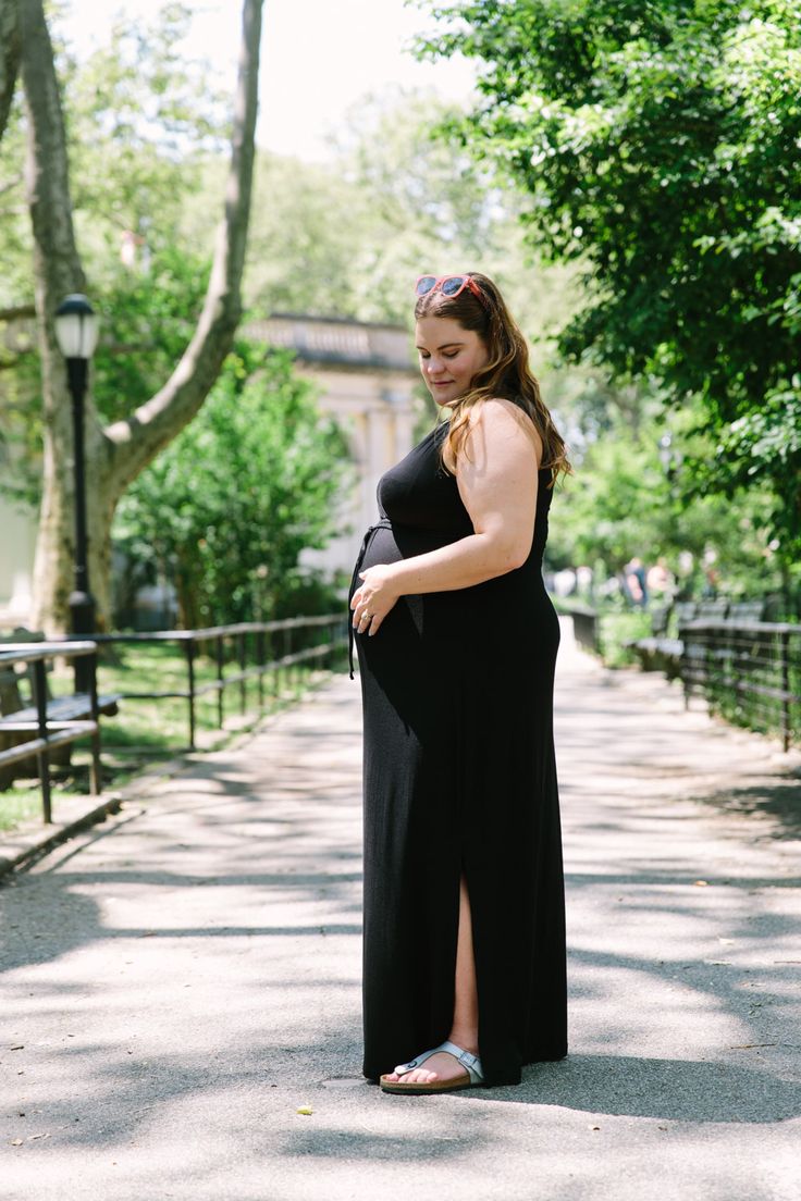 a pregnant woman in a long black dress standing on the sidewalk with trees and bushes behind her