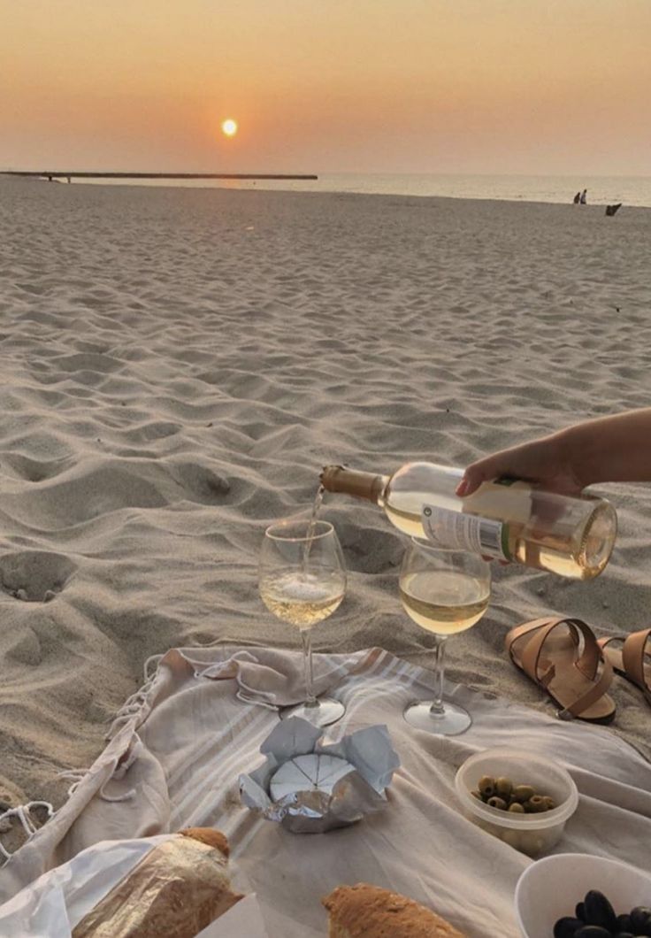 a woman is sitting on the beach with wine and food in front of her as the sun sets