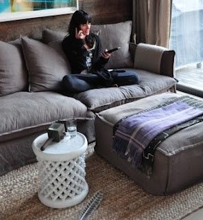 a woman sitting on top of a gray couch in front of a window next to a footstool