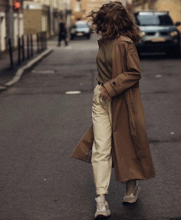 a woman is walking down the street with her hair blowing in the wind while wearing white pants and a trench coat