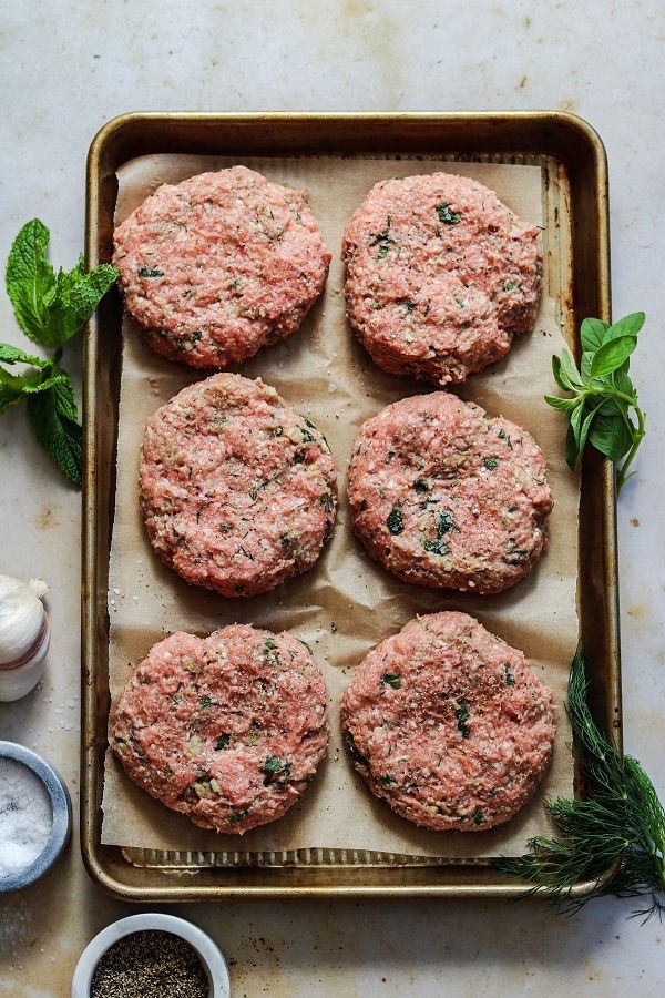 six hamburger patties on a baking sheet with herbs and seasonings next to them