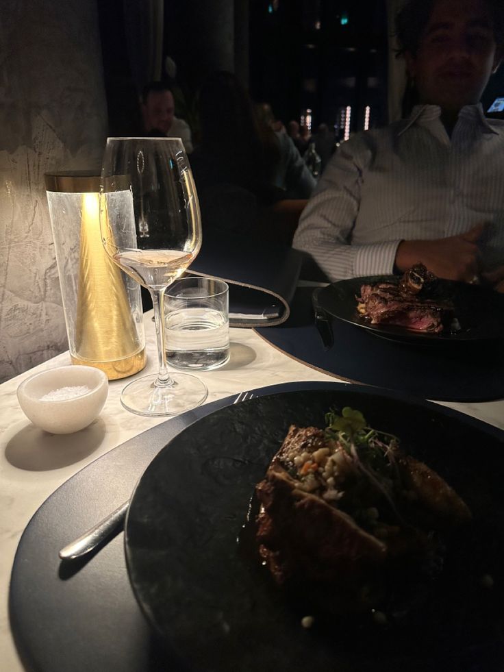 two plates with food and wine on a table in a dimly lit room, next to a man sitting at a dinner table