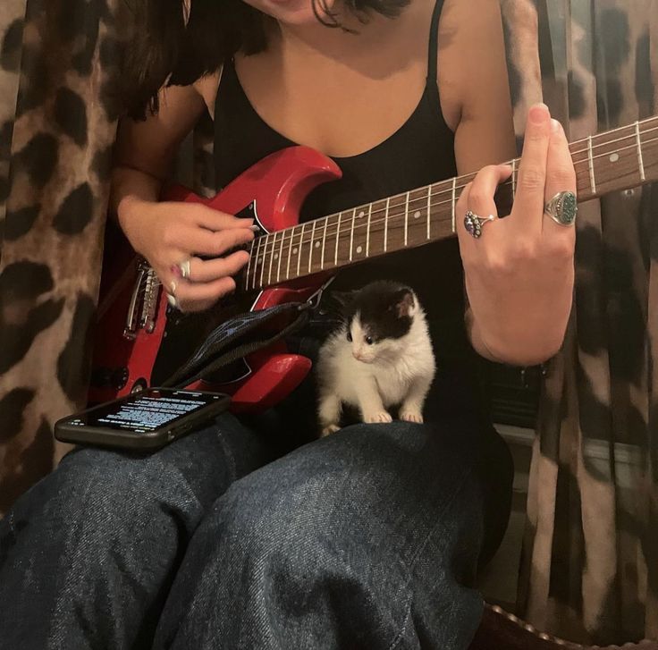 a woman playing guitar with a cat sitting on her lap next to her and holding a red electric guitar