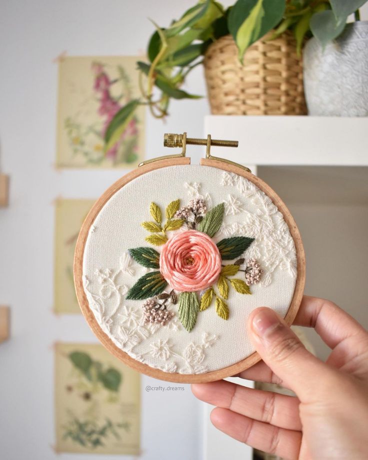 a person holding up a small embroidered flower on a wooden hoop with flowers in the background