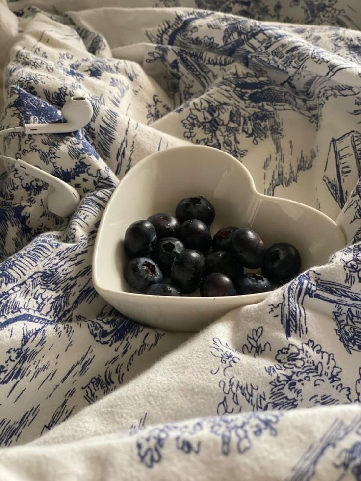 a white bowl filled with black berries on top of a bed covered in blue and white sheets