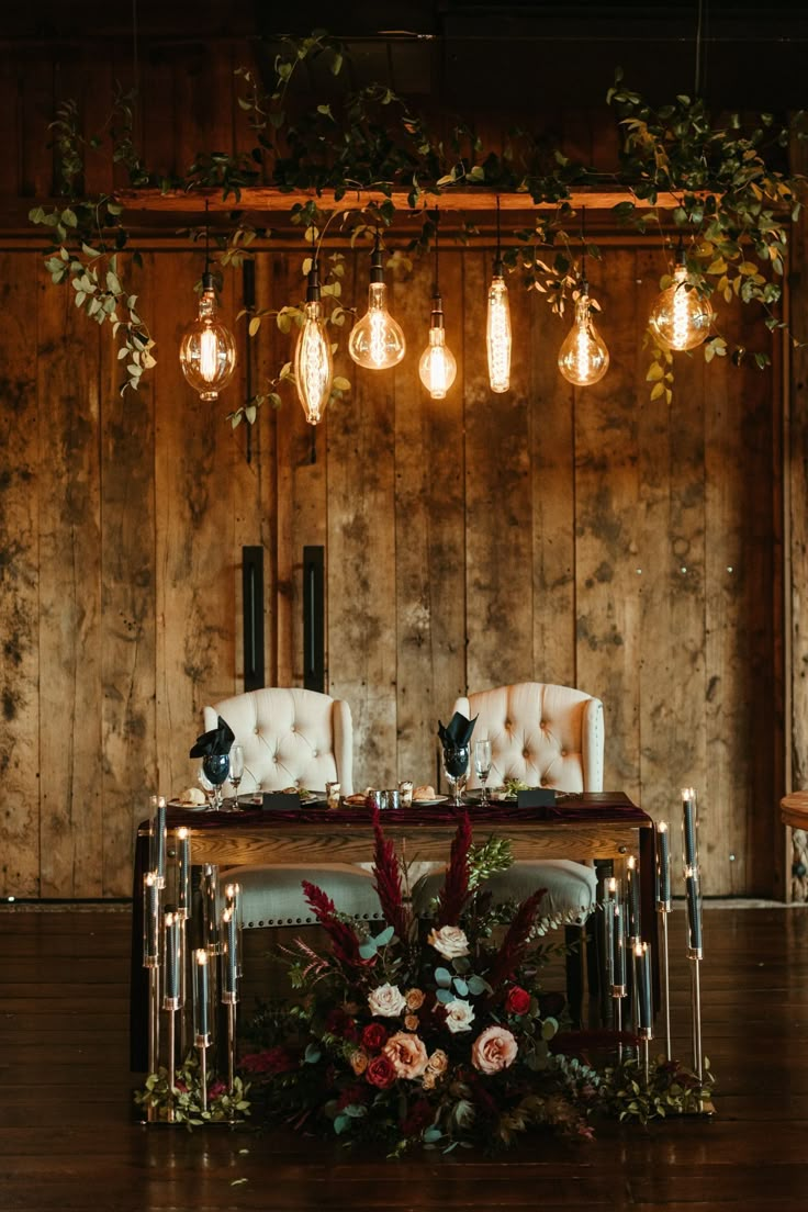 two white chairs sitting at a table with flowers and greenery on it, surrounded by hanging lights