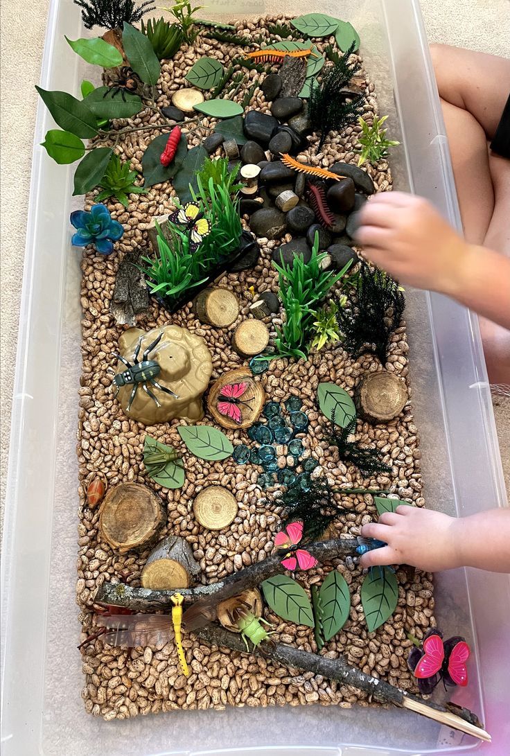 a plastic container filled with lots of different types of plants and rocks next to a child's hand