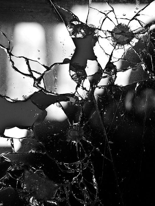 black and white photograph of a person looking through a window with shattered glass on it