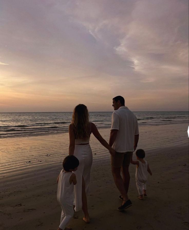 a man and woman holding hands while walking with two children on the beach at sunset