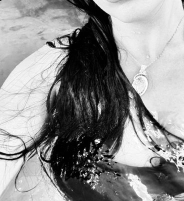 a woman with long black hair is in the water wearing a necklace and looking at the camera