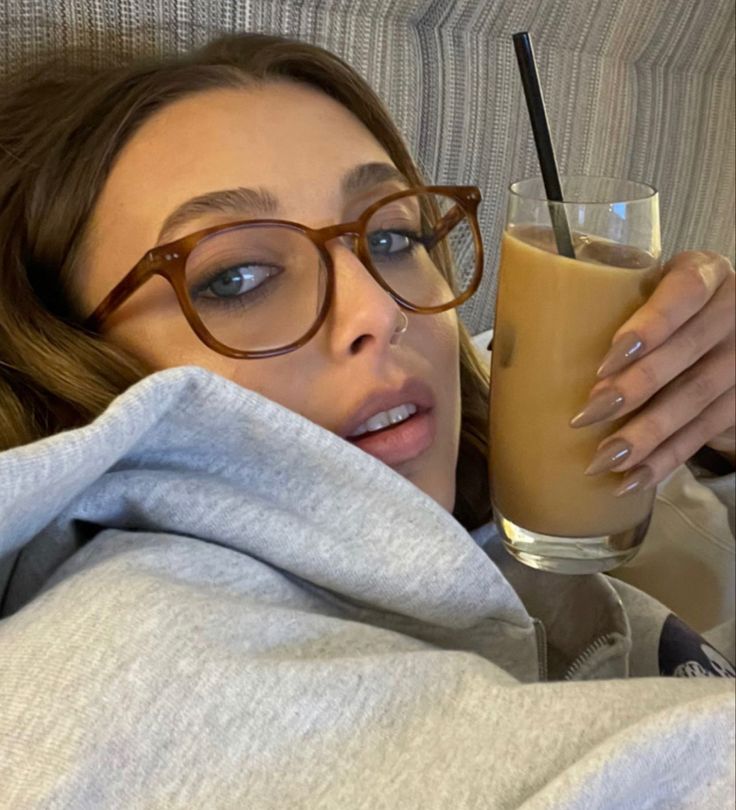 a woman with glasses holding a drink in her hand and looking at the camera while laying on a couch