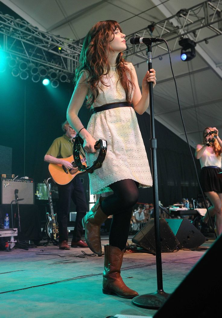 a woman in white dress holding a guitar and singing on stage with other people behind her