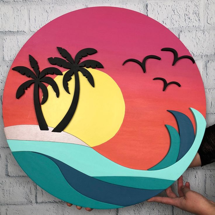 a hand is holding up a painted wooden sign with palm trees and birds on it