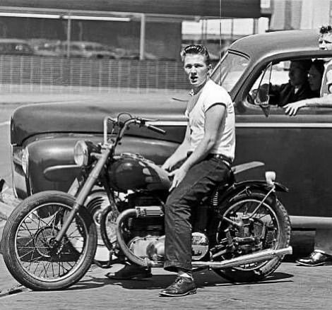 two men sitting on a motorcycle next to an old car