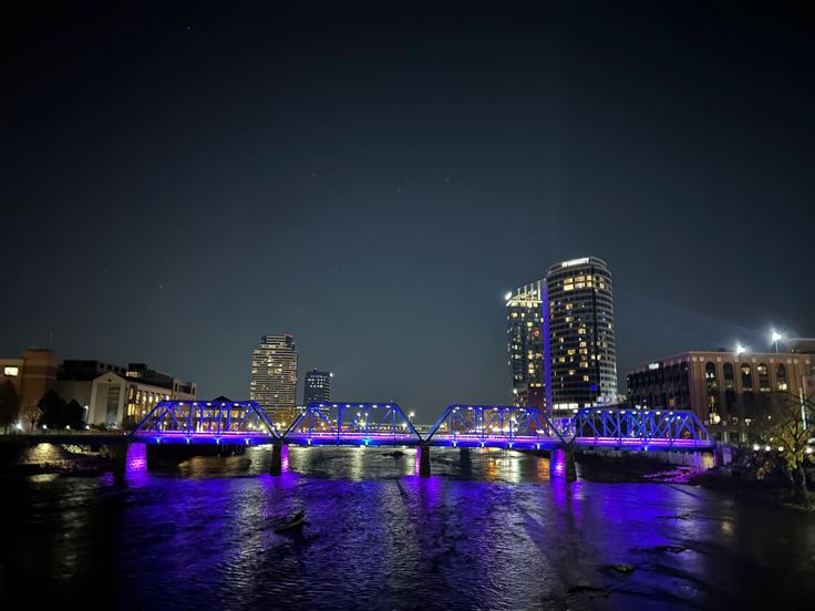 a bridge that is over some water with buildings in the backgroung and blue lights on it