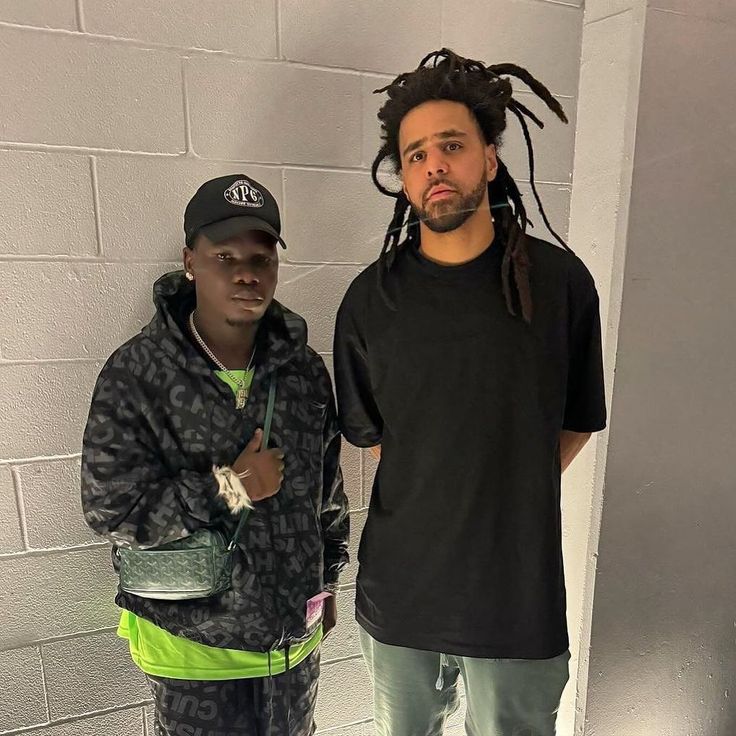 two men standing next to each other in front of a brick wall with dreadlocks on