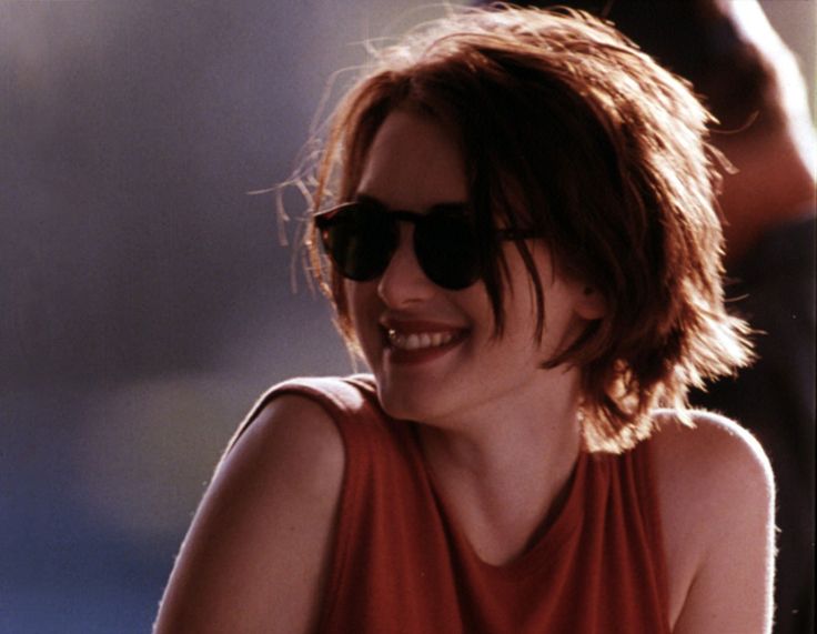 a woman wearing sunglasses sitting down and smiling at someone in the background with her hand on her hip