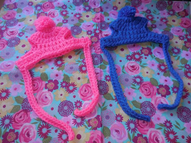 two crocheted hats laying on top of a flowery tablecloth with pink, blue and purple flowers