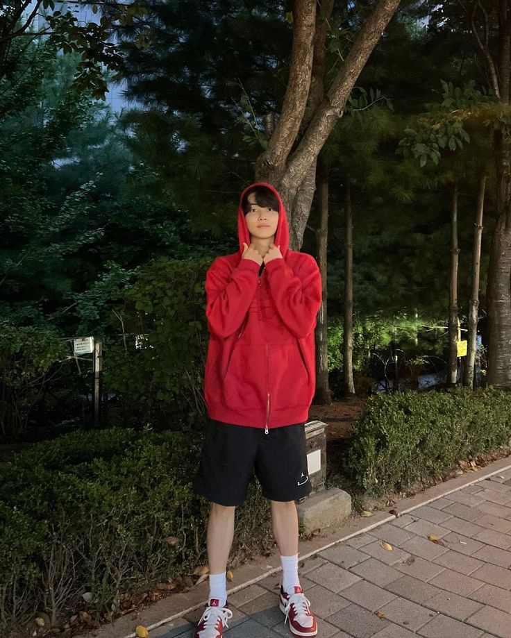 a young man in a red hoodie is standing on the sidewalk with his hands to his face