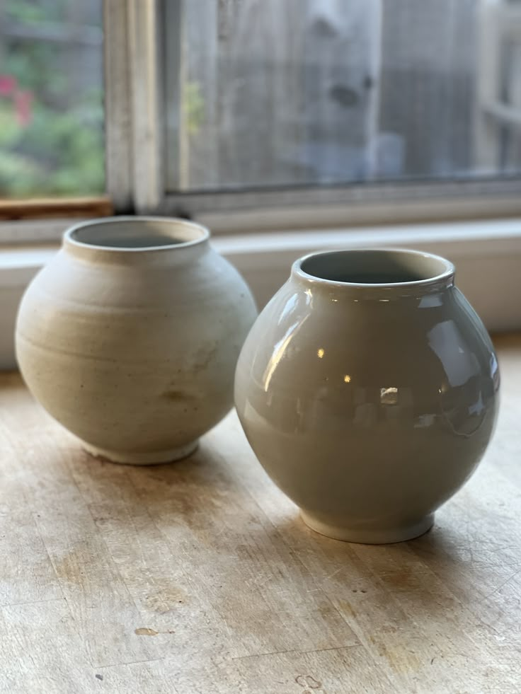 two white vases sitting next to each other on a wooden table in front of a window