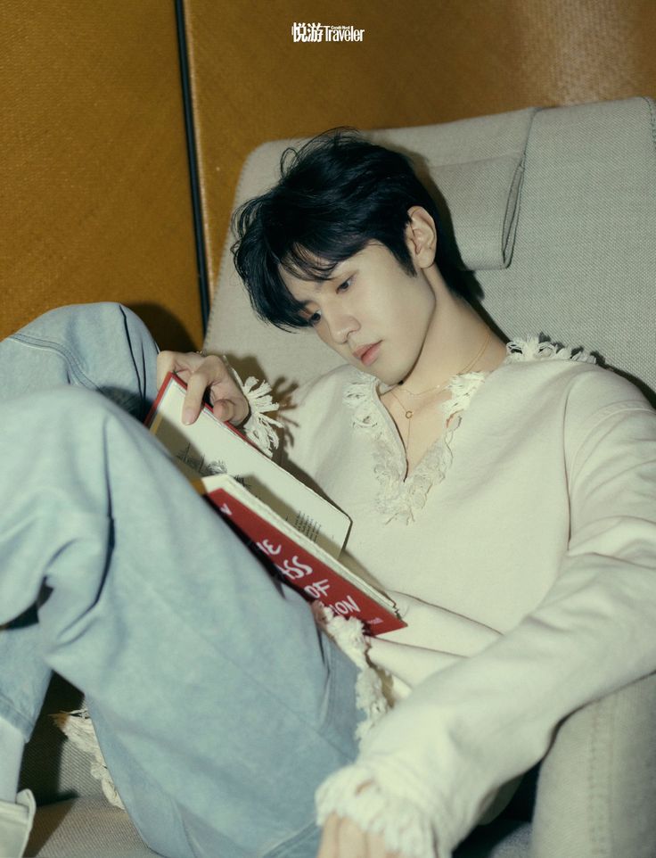 a young man sitting in a chair holding a book and looking down at his cell phone