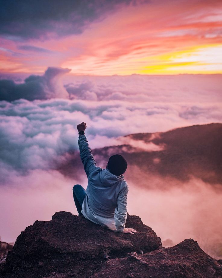 a person sitting on top of a mountain with their arms in the air above clouds