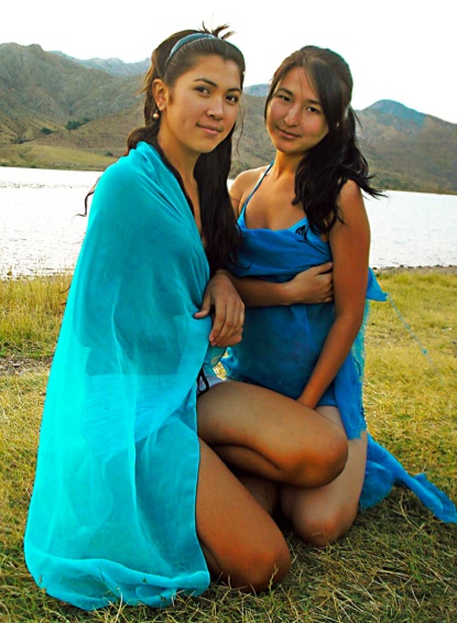 two beautiful young women sitting next to each other on top of a grass covered field