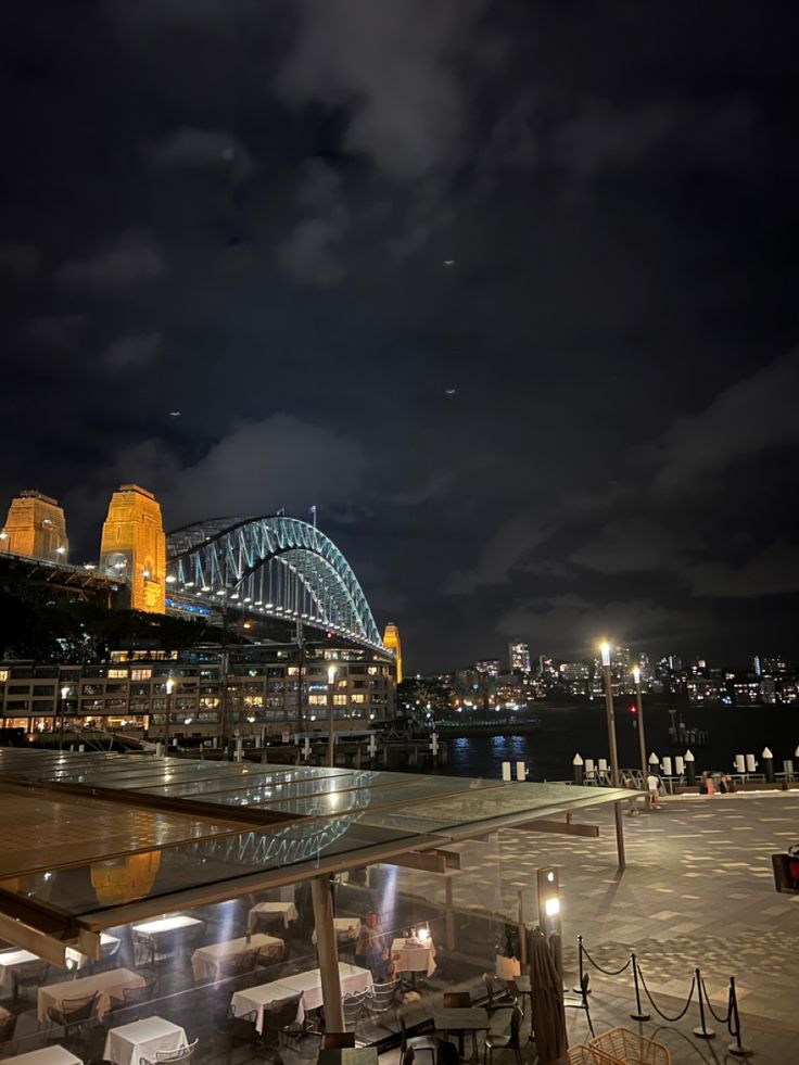 an outdoor dining area at night with the sydney harbour bridge and opera house in the background