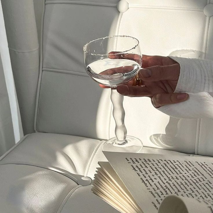 a person is holding a wine glass and an open book on a white leather chair