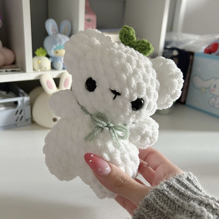 a hand holding a white crocheted teddy bear with a green bow around its neck