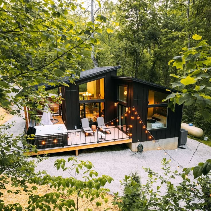 a small black cabin in the middle of some trees with lights on it's deck