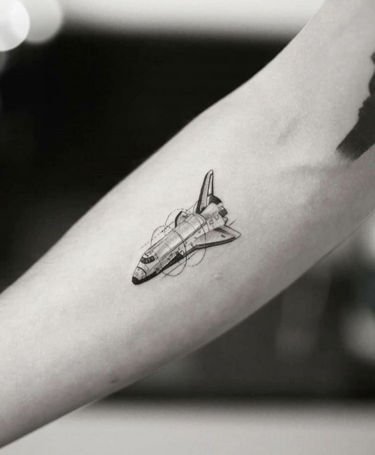 a black and white photo of a small airplane tattoo on the left arm, with an arrow in the middle