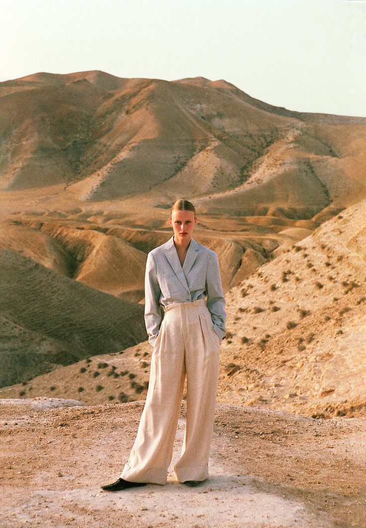 a woman standing on top of a dirt hill next to a desert area with mountains in the background