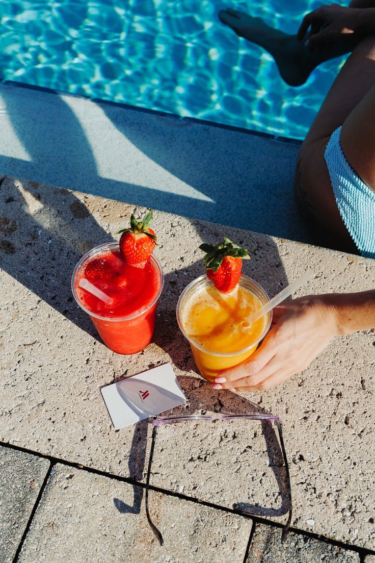 two drinks sitting on the edge of a swimming pool with strawberries and orange juice